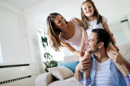 A smiling man sits on a couch with a young girl on his shoulders, while a woman stands beside them in a bright living room.