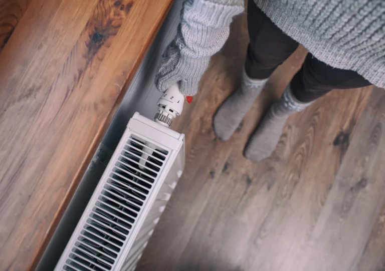 Person wearing a sweater, gloves, and socks adjusts the thermostat on a white radiator next to a wooden surface on a wooden floor.