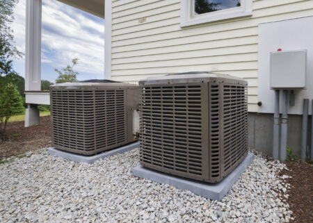 Two outdoor air conditioning units are installed side by side on gravel pads next to the exterior wall of a house.