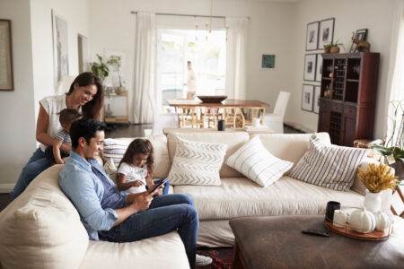 A family of four sits together on a sofa in a bright living room, with one adult standing in the background near the dining area.