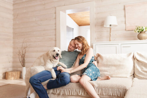 A couple sits closely together on a beige sofa, smiling, while a golden retriever sits beside them in a well-lit, cozy living room.