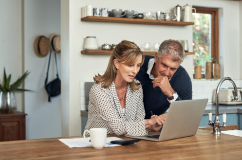 A woman and a man look at a laptop together in a kitchen, with papers and a coffee mug on the counter.
