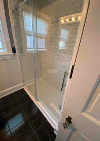A bathroom with a glass-enclosed shower, white subway tile walls, black tile floor, and a window with blinds.