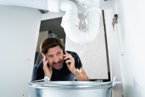 A man on the phone looks at a leaking pipe under a sink, with water dripping into a metal bucket below.