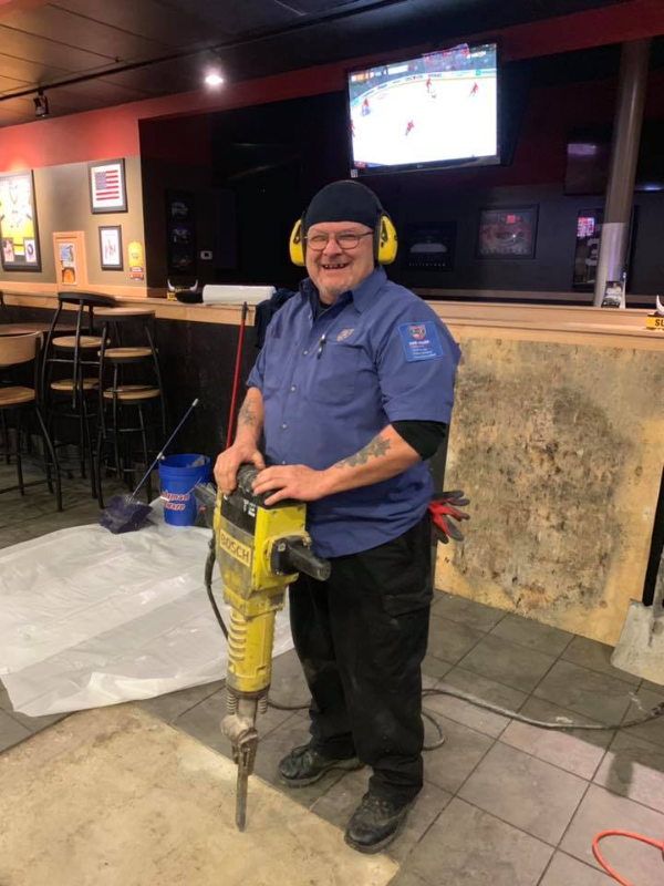 A person wearing ear protection and work clothes operates a large jackhammer inside a restaurant with tables, chairs, and a TV in the background.