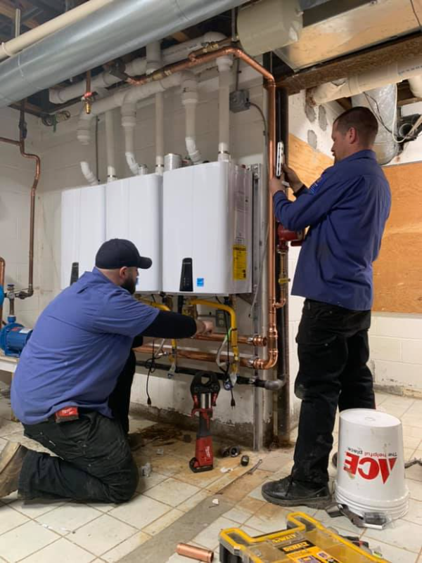 Two workers in blue uniforms install or repair plumbing equipment on a basement wall, surrounded by pipes and tools.