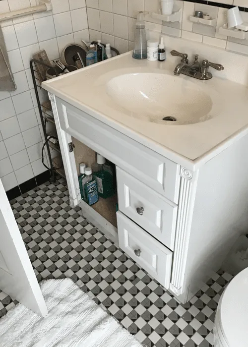 Photo Gallery: White bathroom vanity with an open cabinet door displaying cleaning supplies, two closed drawers, sink, faucet, and toiletry items set on a black-and-white checkered tile floor.