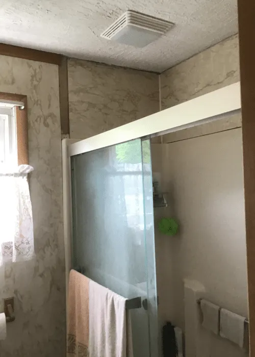 A bathroom featured in the photo gallery with a sliding glass shower door, a towel hanging on the door, a window with a lace curtain, and an exhaust fan on the ceiling.
