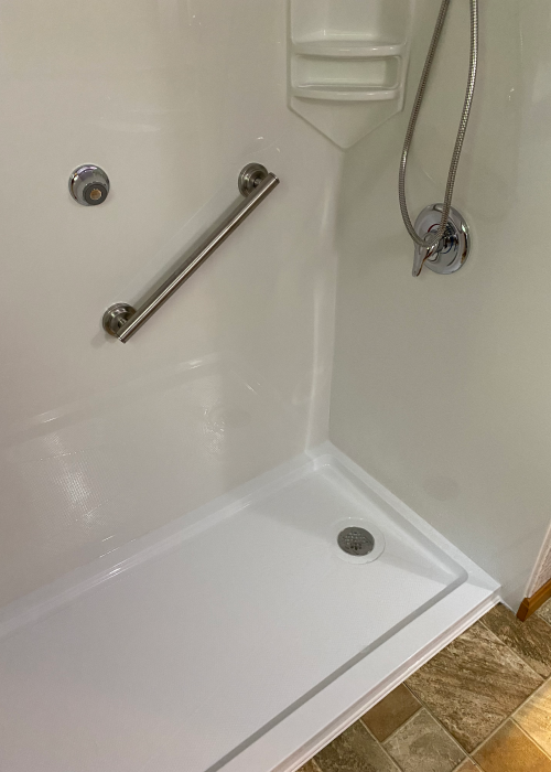 A white shower with a metal grab bar, handheld showerhead, built-in shelf, and floor drain. The shower base meets a tiled bathroom floor—perfect for any modern space featured in a Photo Gallery.