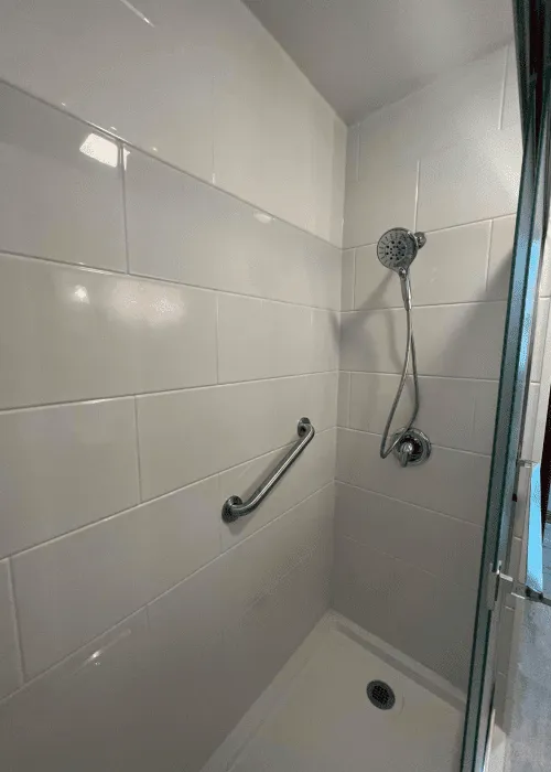 A tiled shower with a detachable showerhead, a horizontal grab bar on the wall, and a drain on the floor—discover more designs in our Photo Gallery.