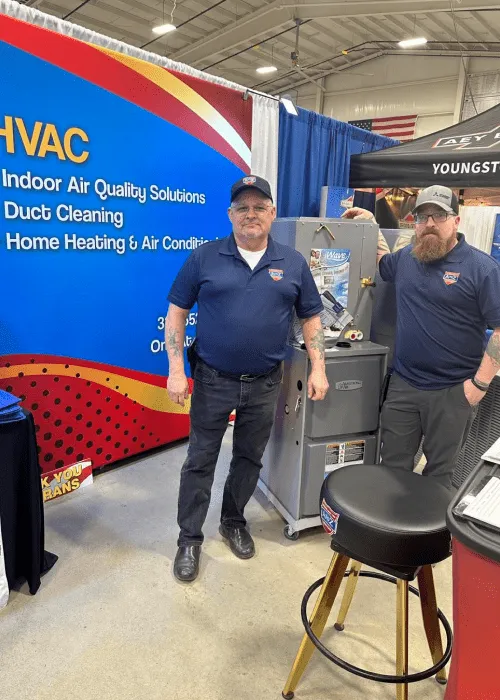 Two men in branded clothing stand beside HVAC equipment at a trade show booth for indoor air quality and heating solutions—see more highlights in our Photo Gallery.