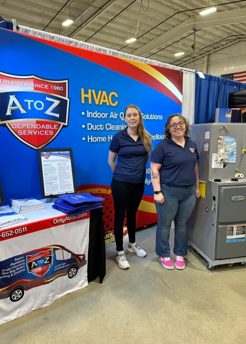 Two women stand at an A to Z Dependable Services HVAC booth with promotional materials and an appliance display in a large indoor event space—perfect for your event’s photo gallery.
