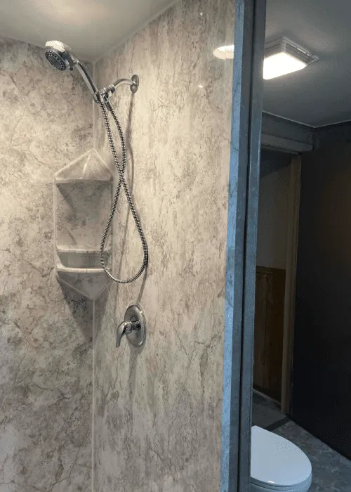 A shower with a handheld showerhead, wall-mounted caddy, and marble-patterned walls is showcased in this Photo Gallery, next to a bathroom with a toilet visible through an open door.