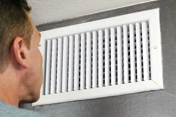 A person looks closely at a white air vent mounted on a gray wall.