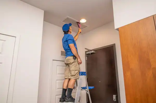 A person standing on a step ladder cleans an air vent on the ceiling inside a residential space.