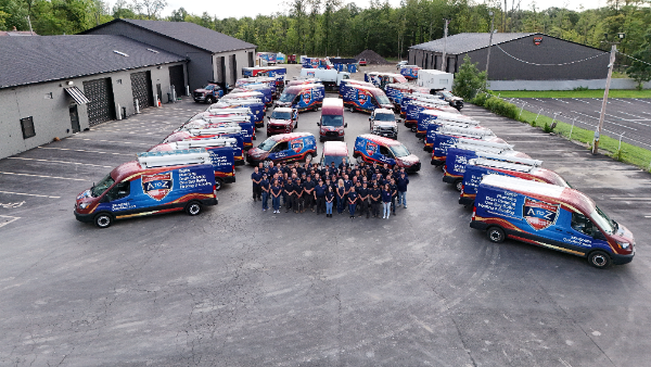 A large group of people stands in front of numerous branded service vans parked in a lot with industrial buildings and trees in the background.