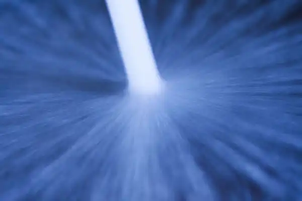 A close-up image of a jet of liquid or gas spraying downward, creating a radial blur pattern against a blue background.