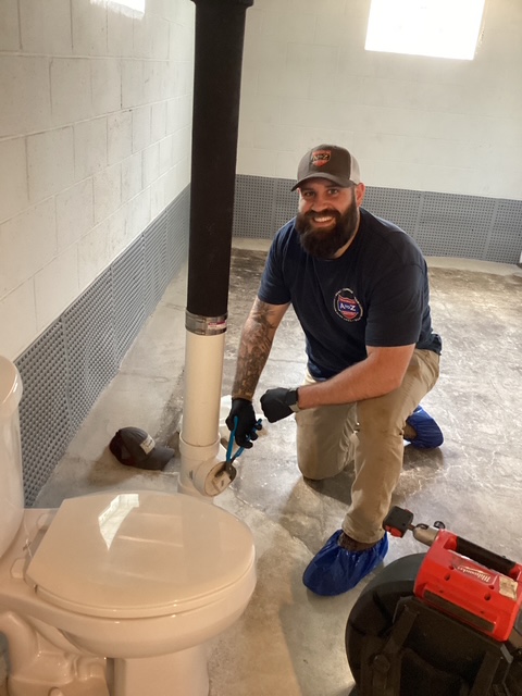 A plumber kneels on the floor next to a toilet, using a pipe wrench on a large vertical pipe in a basement with concrete floors and white block walls.