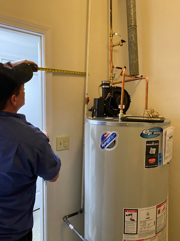 A person in work clothes measures the distance from a wall to a hot water heater with a tape measure in a utility room.