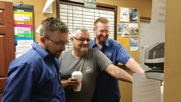 Three men stand together looking at a computer screen in an office, one holding a coffee cup. A wall calendar and documents are visible in the background.
