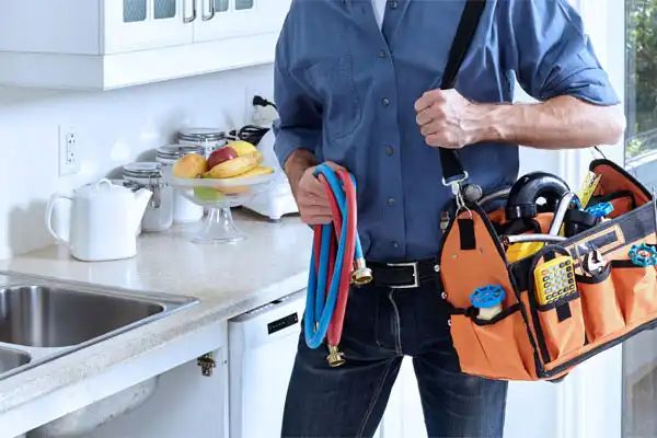 A plumber stands in a kitchen holding blue and red hoses and an orange tool bag filled with plumbing tools.