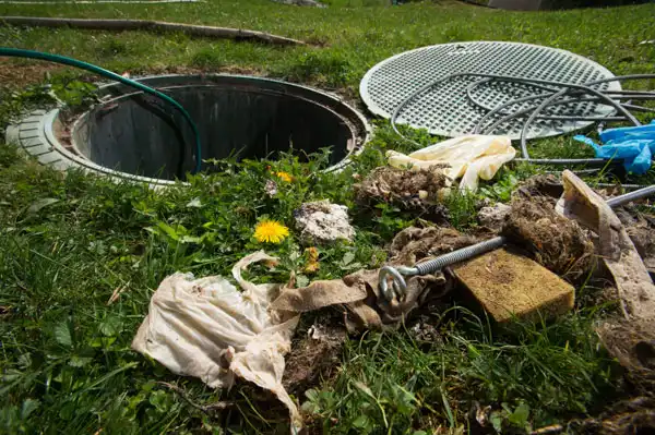 Open septic tank with its lid nearby, surrounded by scattered trash, gloves, debris, and tools on a grassy area.