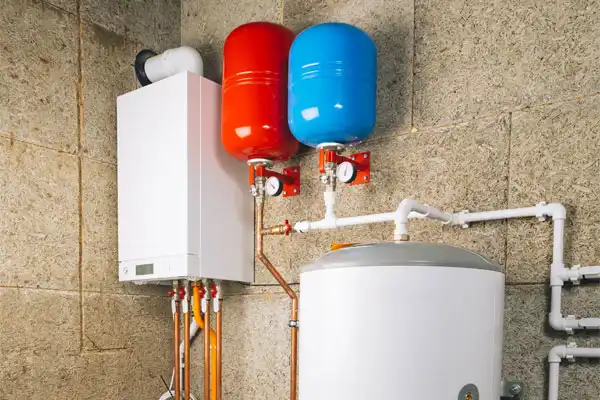 A home boiler system with a white tank, connected pipes, and two wall-mounted expansion tanks—one red and one blue—against a tiled wall.