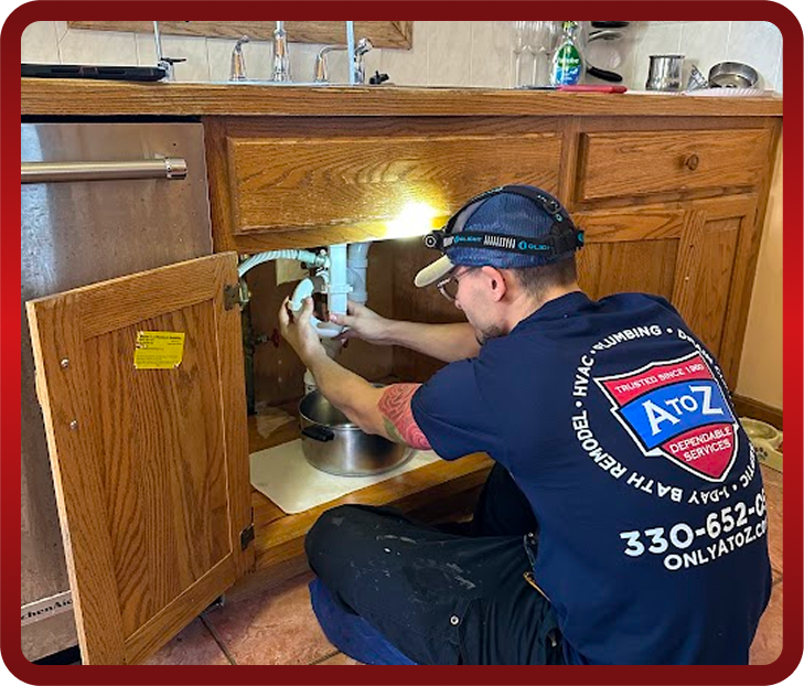 A plumber wearing a headlamp is kneeling and working under a kitchen sink with cabinet doors open, fixing plumbing pipes.