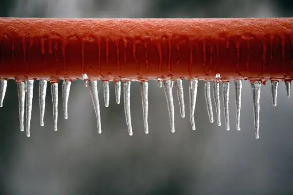 A row of icicles hanging from a red pipe, with a blurred grey background.