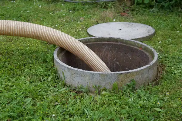 A hose is inserted into an open concrete septic tank on a grassy area, with the round lid set aside.