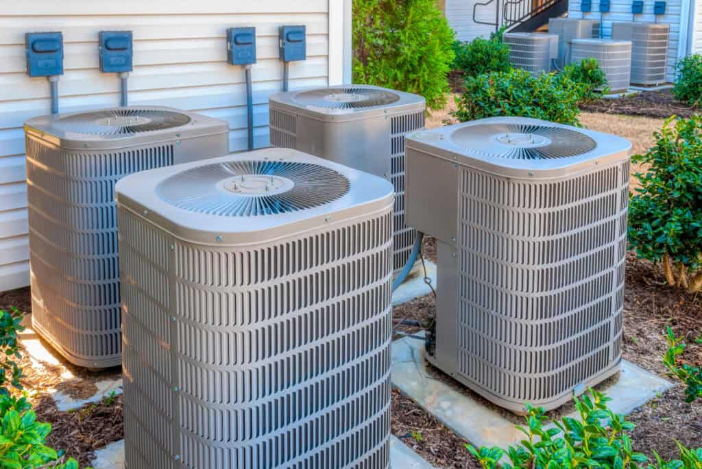 Four outdoor air conditioning units, installed next to the exterior wall of a building, are surrounded by plants and electrical boxes—showcasing the results of professional AC installation and air conditioning services.